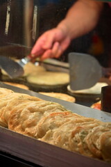 Layered golden-brown scallion pancake with crispy texture in metal tray, chef cooking more on hot griddle behind, famous Xiong Ji Bib Gourmand stall near Gongguan MRT, Taipei.