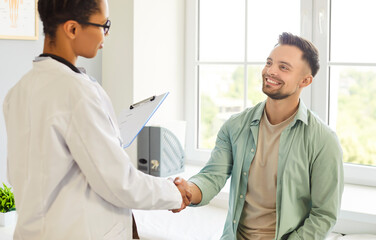 Obraz premium Smiling friendly happy female doctor therapist in white uniform shaking hands with a young patient man standing in office after medical examination in clinic. Medicine and health care concept.