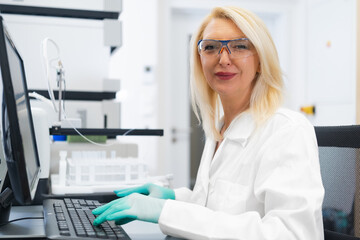 Blonde woman scientist in laboratory working on computer in front of high performance liquid chromatography HPLC equipment
