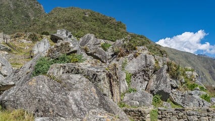 A picturesque mountain against a blue sky and clouds. Dilapidated stone walls, footpaths, and stairs are visible among the mossy boulders. Green vegetation on the slopes. Peru. Machu Picchu.