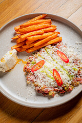 A plate with steak tartare, topped with cheese, chili peppers, and fries, sits on a wooden table