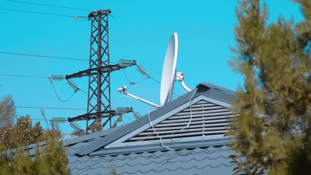 White satellite dish is attached to the roof of a private house against the background of a power line tower on a sunny day. Shot in motion