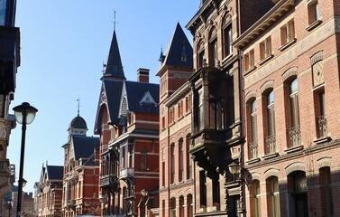 Street view of historic eclectic townhouses in Antwerp, featuring red brick facades, dark slate roofs, and turrets against a clear blue sky.