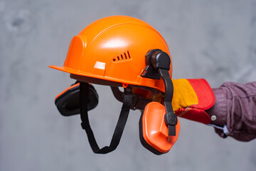 Hand of worker in protective gloves holding orange safety helmet with ear protection. Concept of...