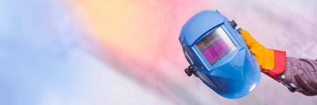 Worker hand in protective gloves holding blue welding helmet on gray concrete background. Concept of industrial safety, personal protective equipment and metalworking tools.