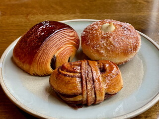 Three traditional Danish baked goods on a ceramic plate. A custard-filled bun, a cinnamon bun, and a chocolate croissant are displayed on a wooden table.