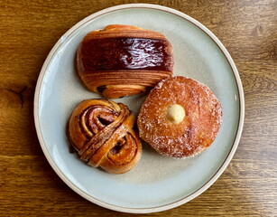 Three traditional Danish baked goods on a ceramic plate. A custard-filled bun, a cinnamon bun, and a chocolate croissant are displayed on a wooden table.