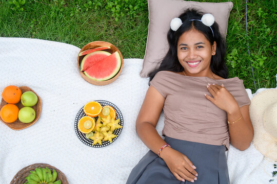 Top view of a smiling young woman lying on a picnic blanket surrounded by tropical fruits and enjoying a sunny outdoor day.