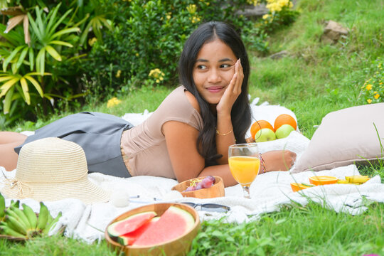 Young woman relaxing outdoors on a picnic blanket with fresh fruits and orange juice, enjoying a peaceful summer day in nature.