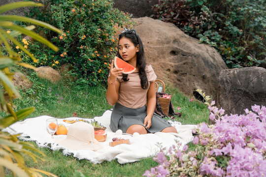 Asian woman enjoying a summer picnic outdoors, holding a slice of watermelon surrounded by flowers and nature.