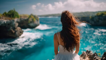 Woman with long brown hair in a white dress stands on a rocky cliff, looking out over turquoise ocean as waves crash below. Concept Coastal cliff portrait, White dress, Long brown hair
