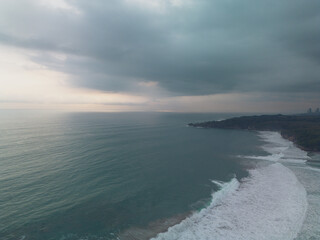 Aerial view of a dramatic coastline with dark clouds, ocean waves, and a distant city skyline at dusk or dawn