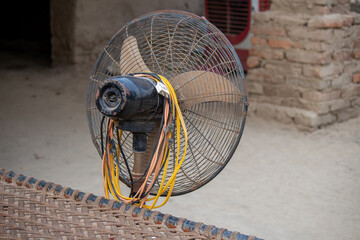 Old electric pedestal fan with worn metal blades in rural village background outdoor scene