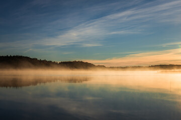 Golden sunrise over calm autumn sea with forest on the shore and gentle fog above the water, peaceful natural seascape and morning atmosphere