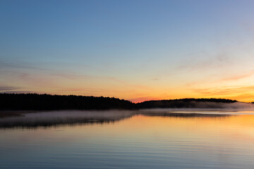Golden sunrise over calm autumn sea with forest on the shore and gentle fog above the water, peaceful natural seascape and morning atmosphere