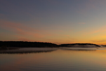 Golden sunrise over calm autumn sea with forest on the shore and gentle fog above the water, peaceful natural seascape and morning atmosphere