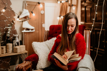 Young woman in a cozy red sweater sitting in a plush armchair, relaxing and reading a book in a...