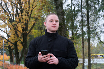 A guy in a black jacket stands against the background of an urban autumn landscape with a phone in his hand