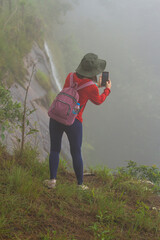 A woman standing at the edge of a cliff is about to take a picture of a waterfall from the cliff with her mobile phone.