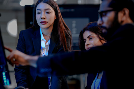 Two diverse professionals in suits collaborate in a modern tech office. One uses a stylus on a tablet while the other observes, analyzing complex data on multiple screens.