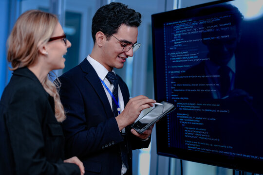 Intense code walkthrough. A female developer uses a stylus pointing to a specific line of code on the screen explaining its function to her male teammate during a debugging session.