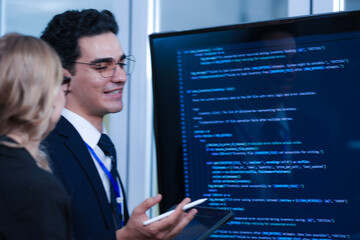 Dynamic duo collaborates late night. Focused male and female professionals discuss project details reviewing info on tablet and computer screen in modern tech workspace.