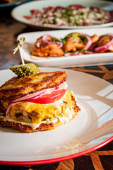 A colorful layered sandwich features cheese, veggies, and a pickle on a beautifully designed plate