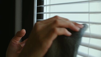 Hand meticulously wiping dust from white horizontal window blinds with soft gray cloth, showcasing routine domestic chore and general housekeeping task. Person cleaning white window blinds with cloth