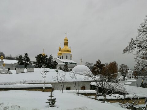  Kyiv Pechersk Lavra, located in Ukraine, is shown in a winter landscape. The scene features snow-covered roofs and trees, with the prominent golden domes and crosses  - Powered by Adobe