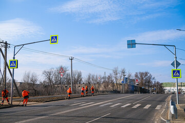 Workers in orange overalls are cleaning the side of an urban or suburban road on a spring day.