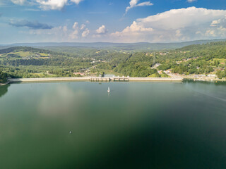 A scenic view of Solina Dam, Poland, surrounded by lush greenery and hills under a bright sky. A sailboat glides on the tranquil water, showcasing natures beauty.