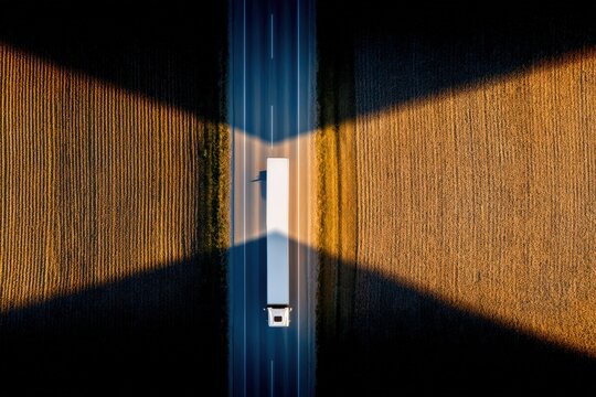 Aerial perspective of a freight truck driving along a highway bordered by golden fields casting dramatic shadows highlighting the intersection of industry and agriculture