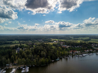 Obraz premium Aerial view of Mikolajki, Poland, showcasing a vibrant landscape with lush green trees, a calm lake, and numerous boats docked along the shoreline under a vast sky with clouds.