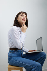 A thoughtful young woman sits on a stool with a laptop, gazing upward as she ponders ideas in a clean, minimalist setting that highlights focus and modern remote work.