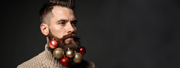 A man in a beige sweater with a neat beard decorated with Christmas balls on a dark gray background