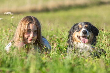 Smiling Australian Shepherd dog sitting beside a young girl in a sunny field. The relaxed outdoor portrait captures a moment of friendship, warmth, and natural connection between human and animal.