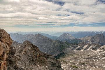 Impressive view of the surrounding mountain peaks near Zugspitze in summer. Rugged rocks, snow patches, and distant valleys form a striking alpine landscape under a partly cloudy sky.