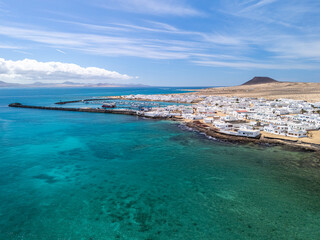 Aerial view of the coastal village on La Graciosa island with white houses, turquoise ocean water, and volcanic landscape in the background.  
