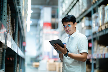 A man wearing glasses is standing in a warehouse with a tablet in his hand. He is focused on the tablet, possibly checking inventory or processing orders