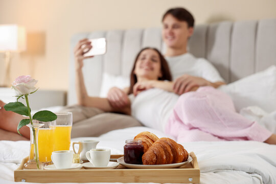 Tray with delicious breakfast on bed and happy couple taking selfie at hotel room, selective focus