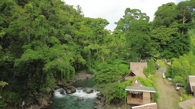 Slow rising drone reveals an emerald river with rapids and boulders beside nipa huts and a leafy path at Tampisaw River & Eco Park, Napsan, Puerto Princesa, Palawan, Philippines.