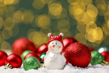 Cute decorative snowman and shiny baubles on artificial snow against blurred lights, closeup