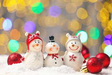 Cute decorative snowmen and red baubles on artificial snow against blurred lights, closeup