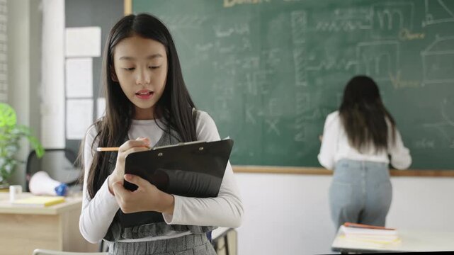 Back to school concept. Portrait of happy Asian child girl student in uniform with school bag and backpack standing holding clipboard with writing and taking notes in front of blackboard in classroom.