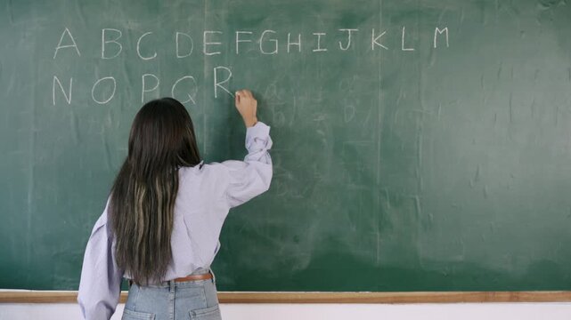 Video English basic class online concept. Happy young Asian female teacher stands writing and teaching English language lesson on the blackboard via video call on a technology smartphone in classroom.