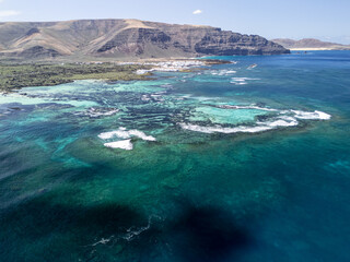 Naklejka premium Aerial view of volcanic reef patterns and turquoise shallow waters near Órzola, Lanzarote, with mountains and coastline in the background. 