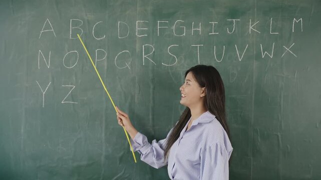 Video English basic online concept. Happy Asian woman teacher standing holding a pointer and teaching English language lesson on the blackboard via video call in the classroom. Back to school concept.