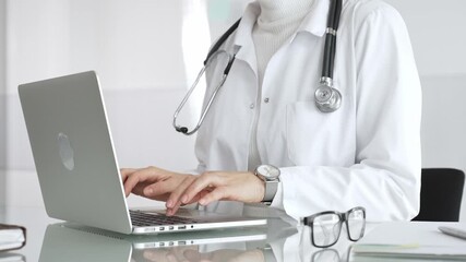 Woman doctor wearing white coat and stethoscope, typing on laptop in clinic office, using modern medical technology. Medicine, healthcare and science concept - Powered by Adobe