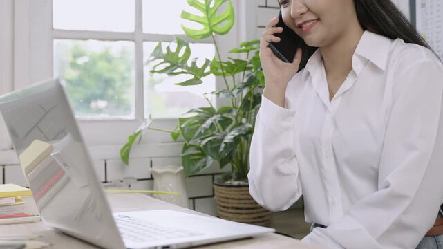 Closeup of happy young Asian female teacher sitting smiling and typing keyboard on laptop computer and using her mobile phone technology talking at her desk in classroom. Back to school concept. 