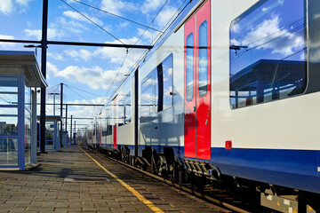 Modern train with red doors at a sunlit platform. Blue sky and clouds reflect vividly in the windows, highlighting the urban railway infrastructure.